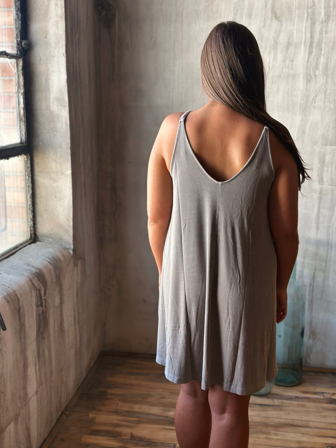 Woman wearing a gray sleeveless dress standing in a room with a window and textured wall.