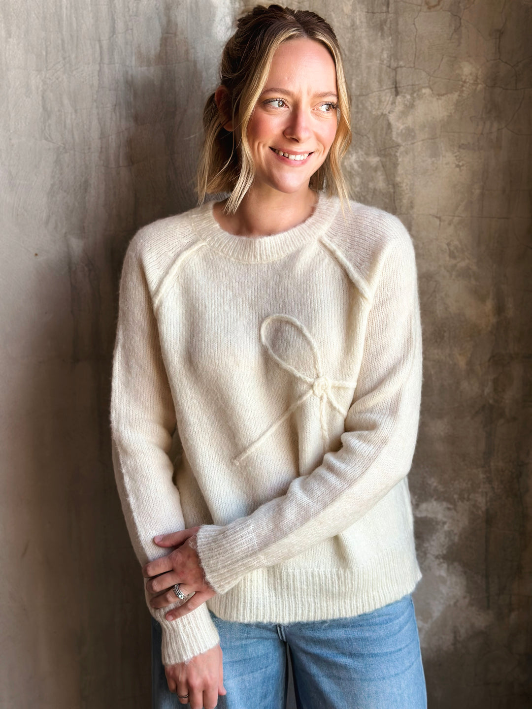 Woman wearing a cream sweater with a heart design against a textured wall.