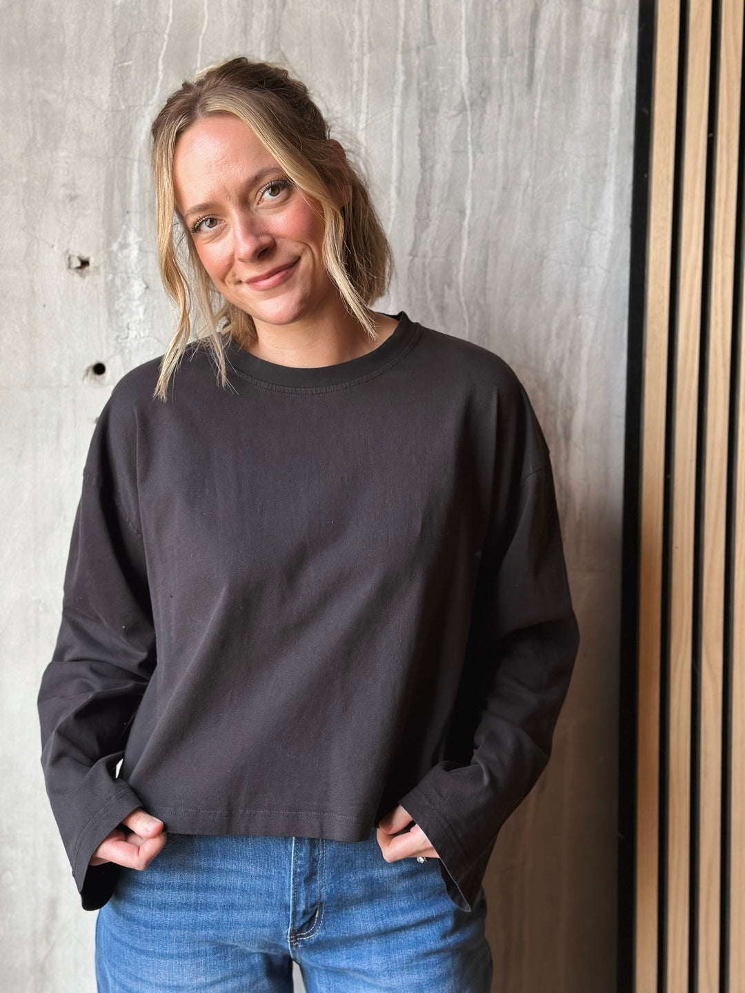 Person wearing a black sweatshirt and blue jeans standing against a textured wall.