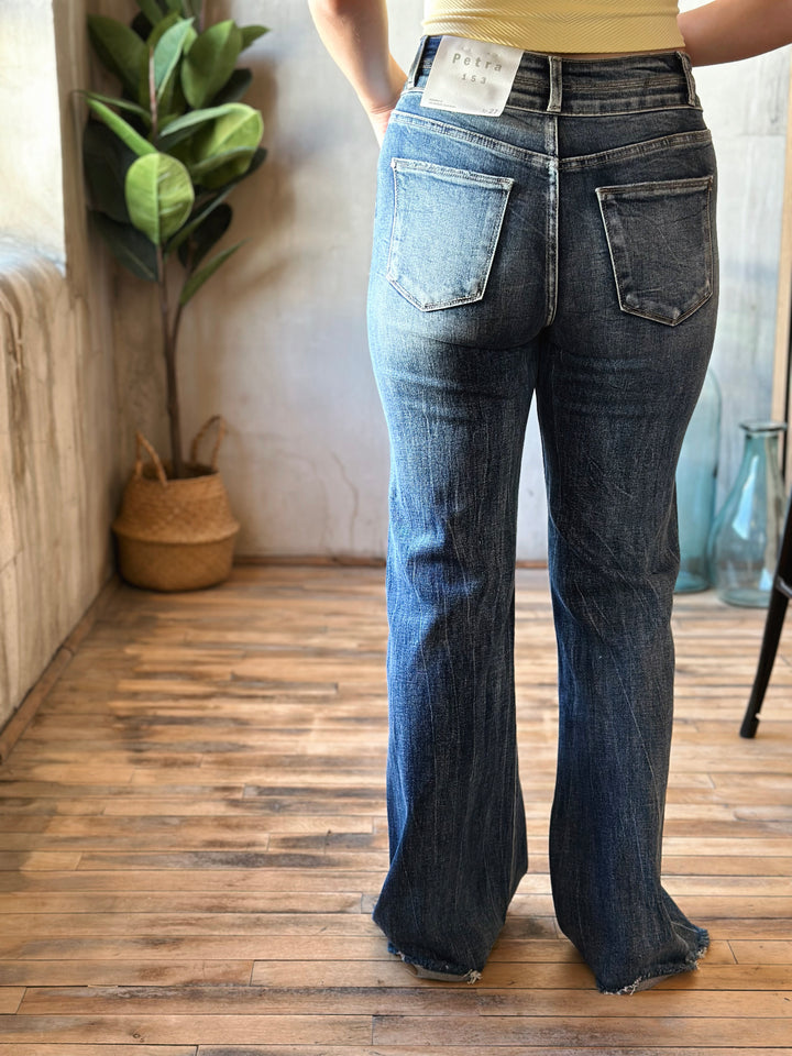 Person wearing blue flared jeans standing on a wooden floor with a plant in the background