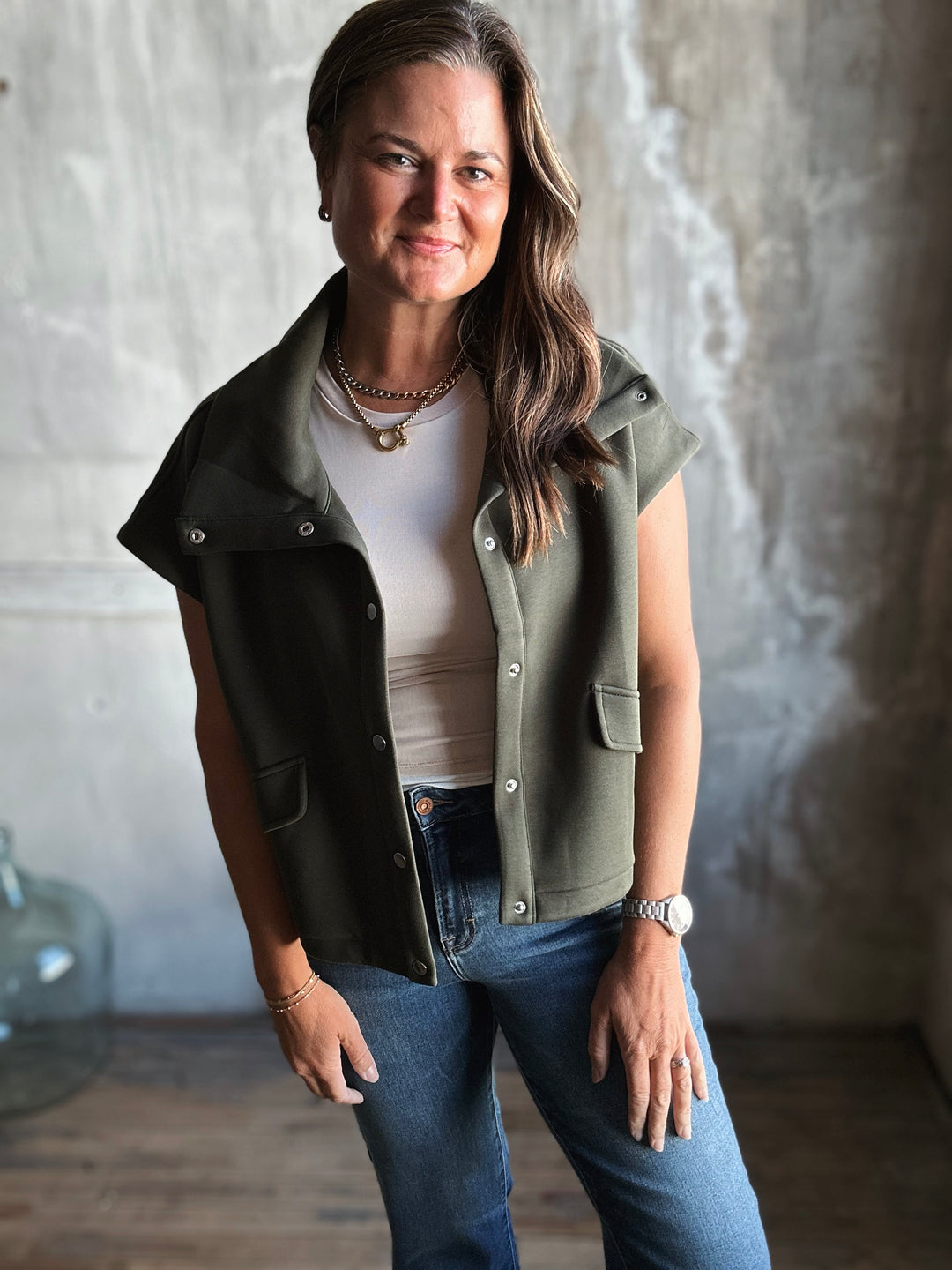Woman wearing a green vest over a white shirt and blue jeans in a neutral setting