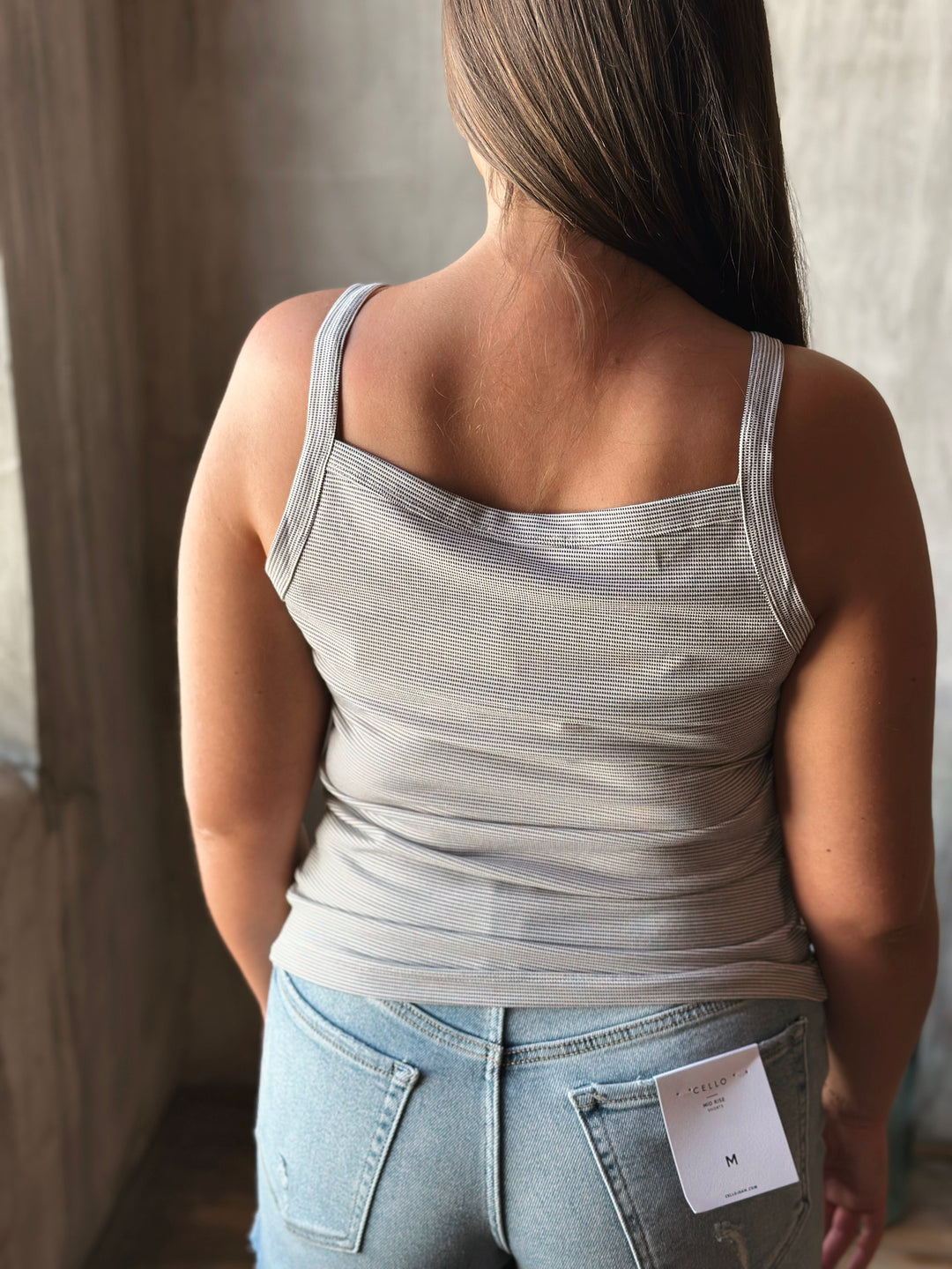 Person wearing a light gray tank top and blue jeans with a tag, standing indoors.