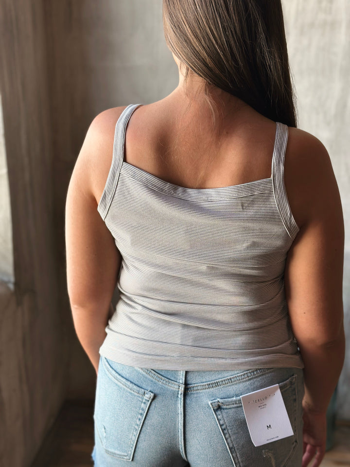 Person wearing a light gray tank top and blue jeans with a tag, standing indoors.