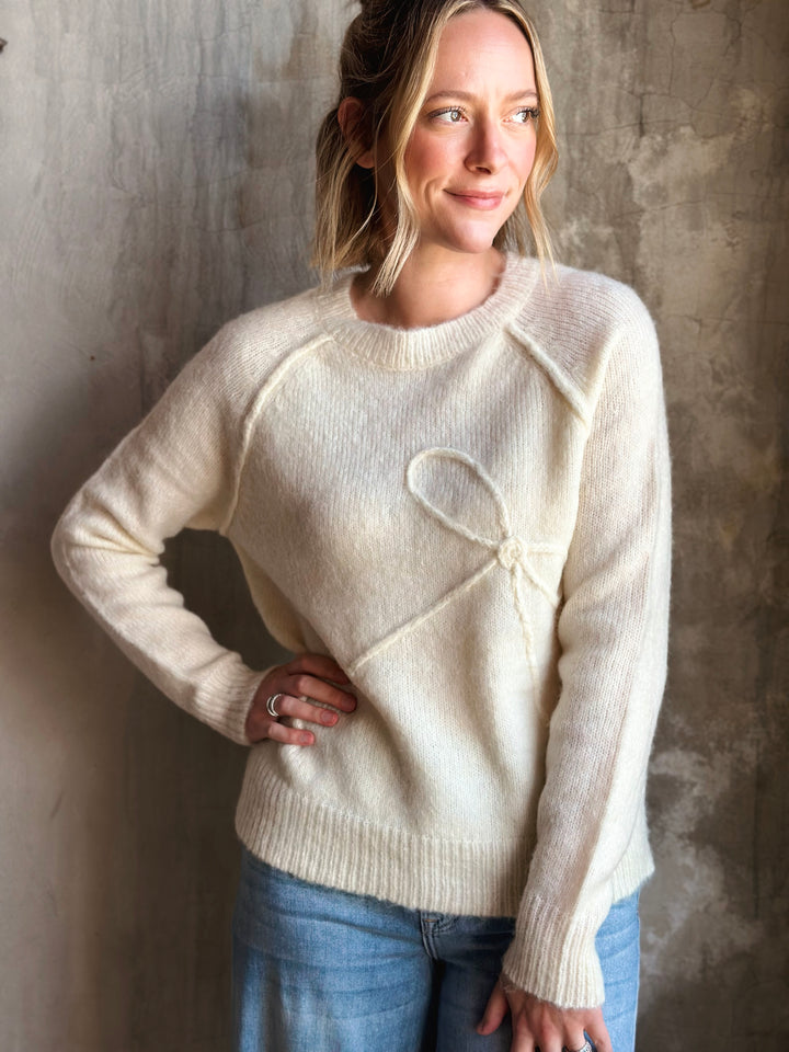 Woman wearing a cream sweater with a heart design against a textured wall.