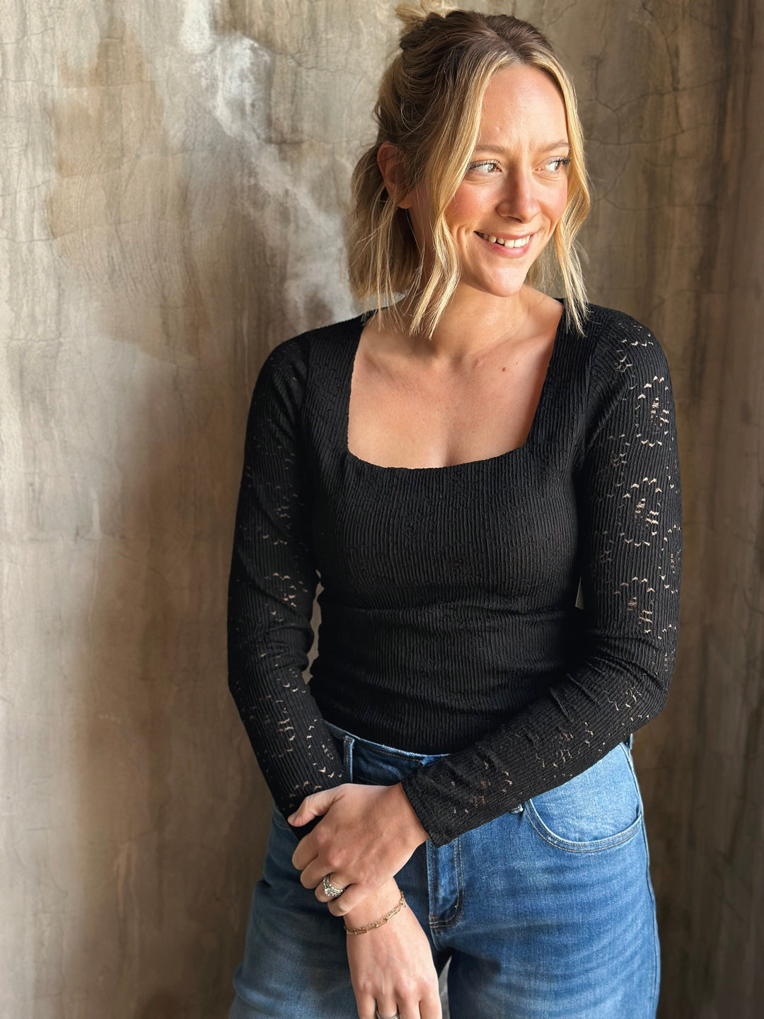 Woman wearing a black long-sleeve top and blue jeans against a textured wall.