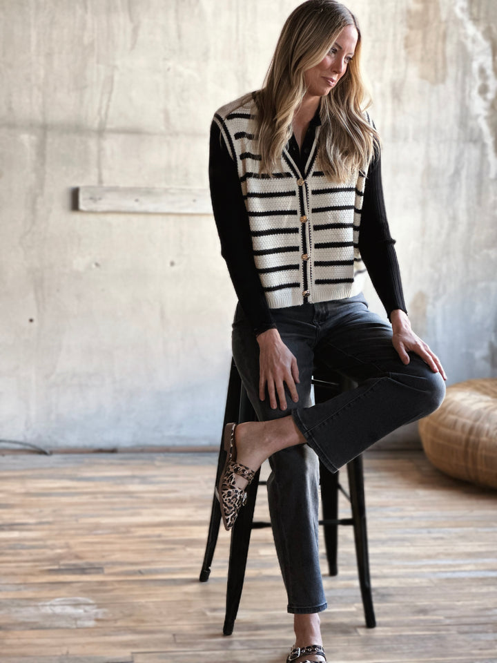 Woman sitting on a stool wearing a black and white striped sweater and black pants in a room with wooden floor and concrete wall.