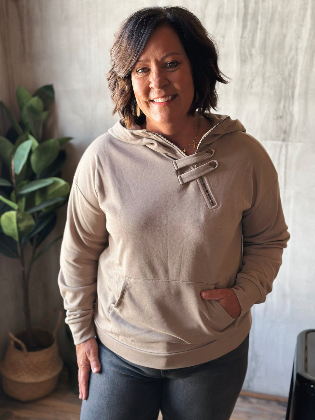 Woman wearing a beige hoodie standing indoors with a plant in the background