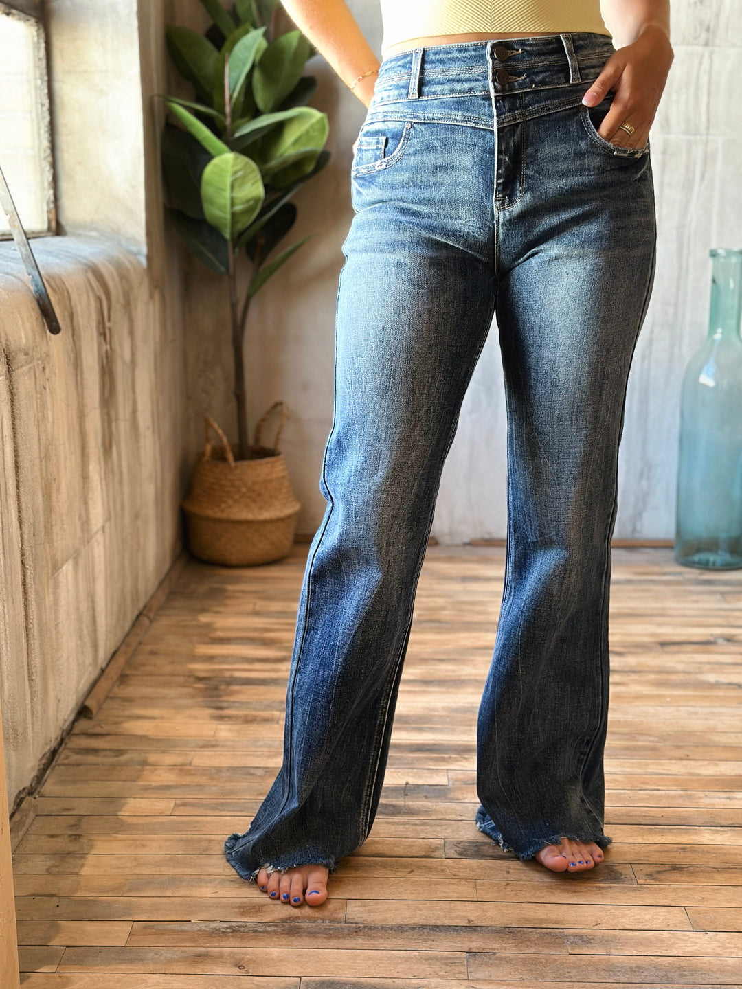 Person wearing blue jeans standing in a room with wooden floor and plant.