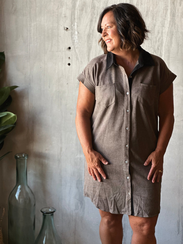 Woman wearing a brown button-up dress standing against a textured wall.
