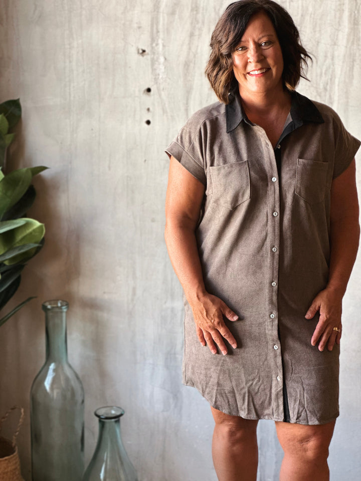 Woman wearing a sleeveless shirt dress standing against a textured wall with decorative bottles and plants.
