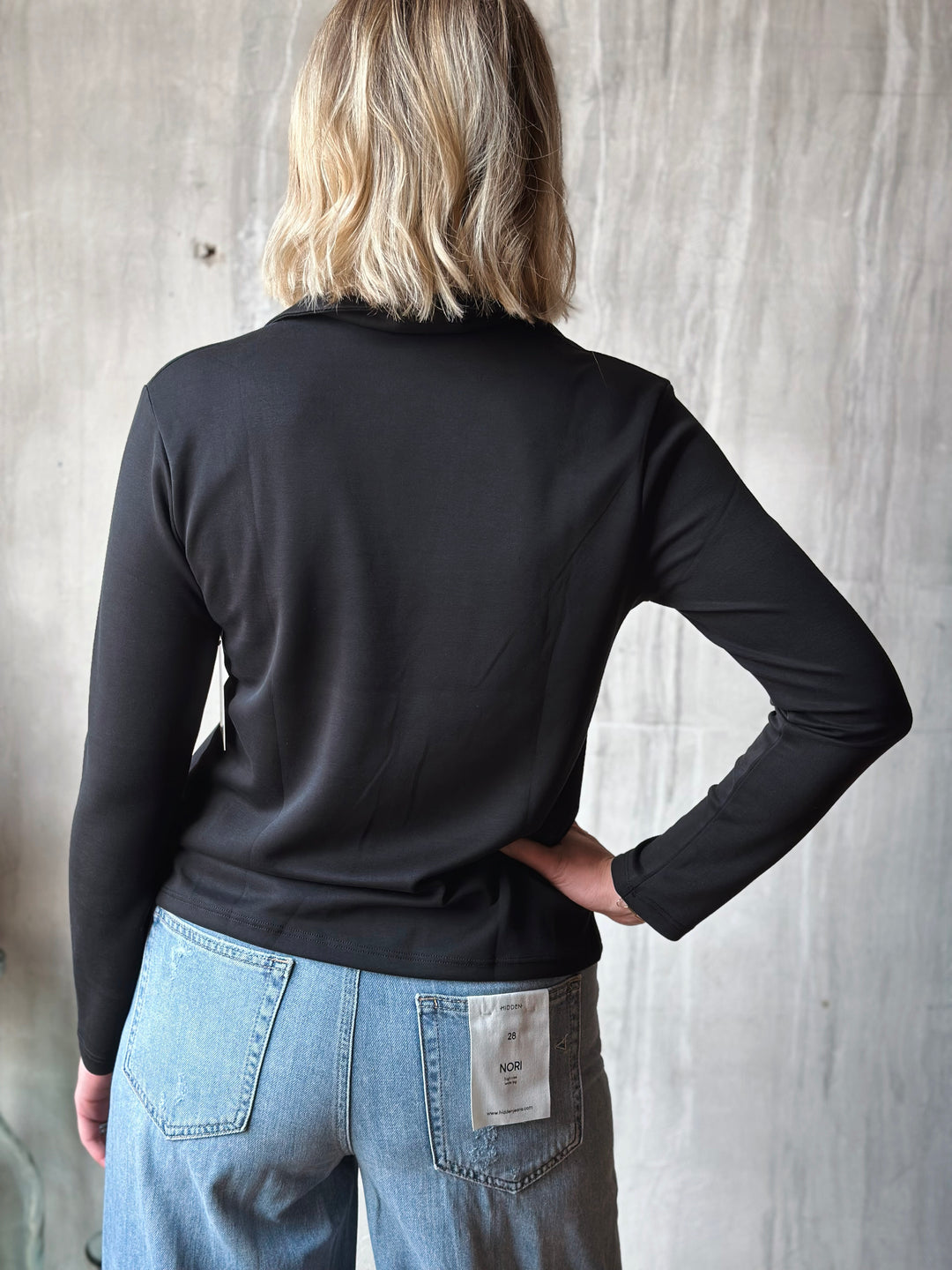 Person wearing a black long-sleeve shirt and blue jeans against a wooden wall.