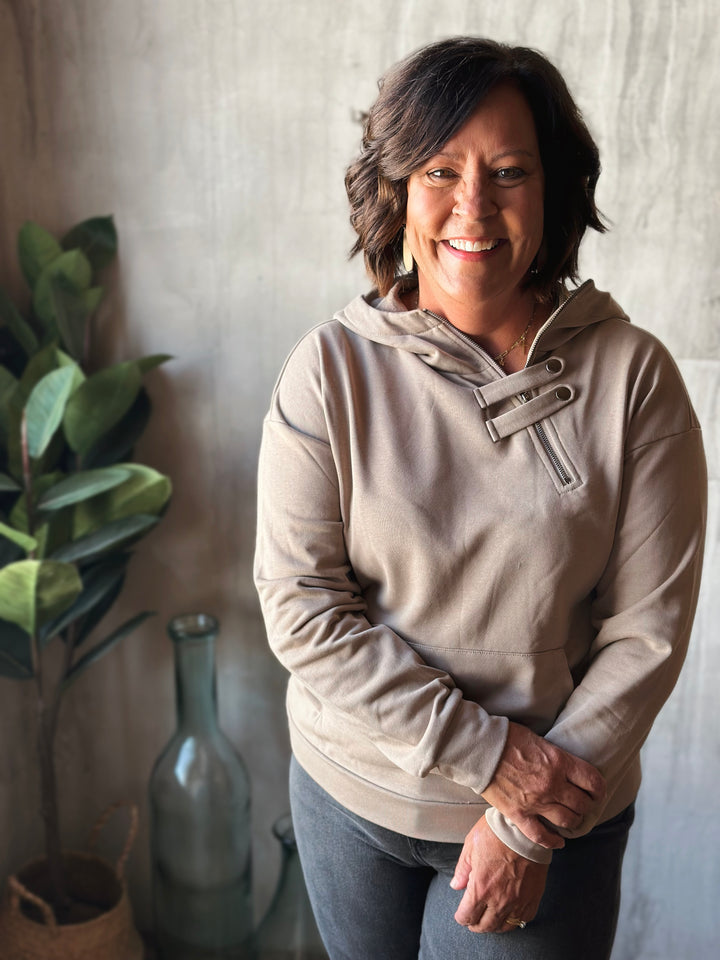 Woman wearing a beige hoodie standing indoors with a neutral background