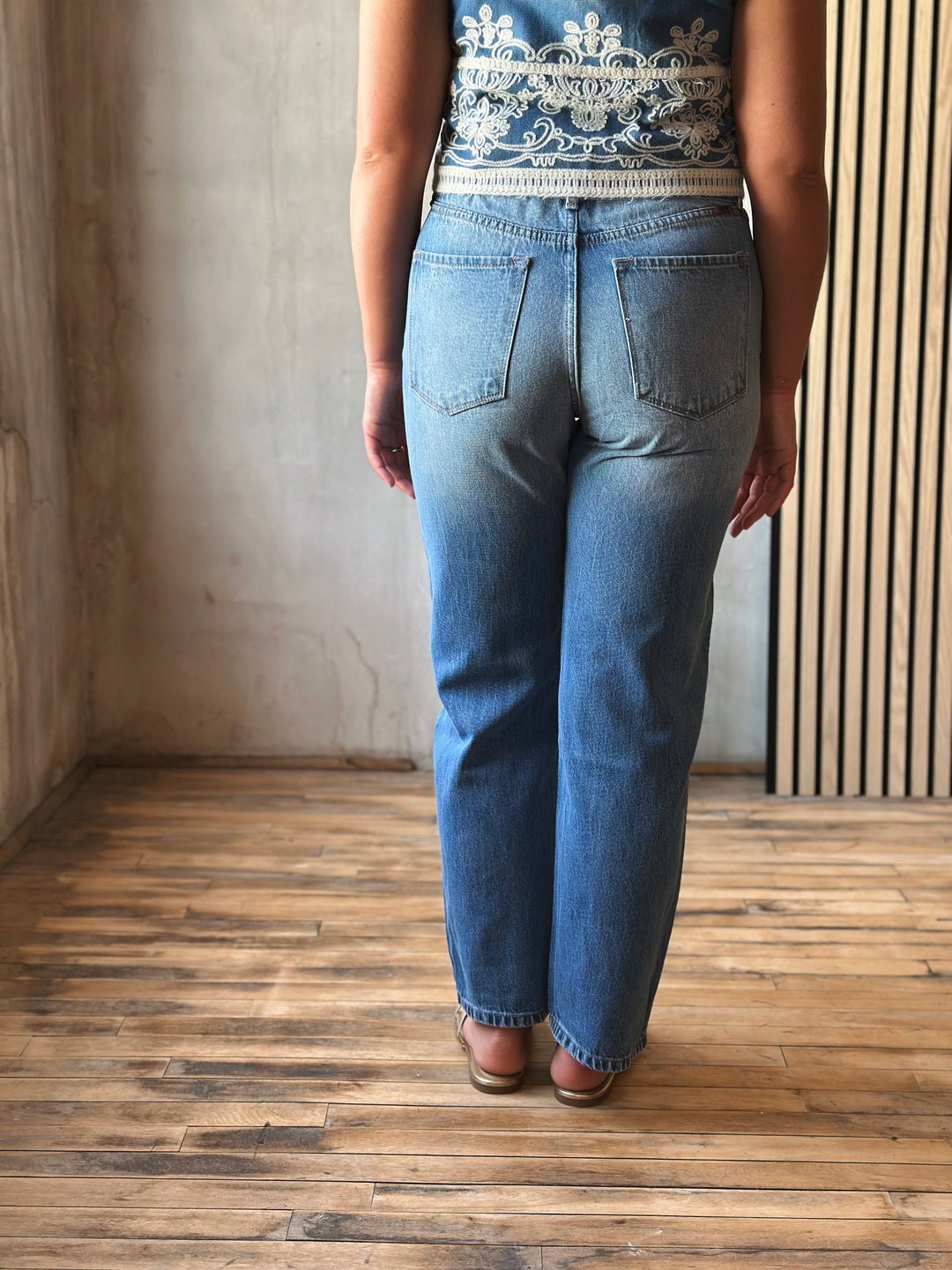Person wearing blue jeans and a patterned top standing on a wooden floor.