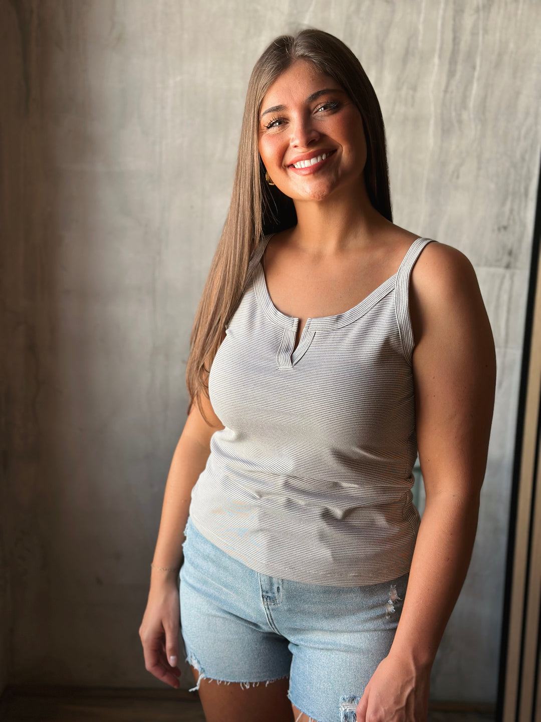 Woman wearing a light gray tank top and denim shorts standing against a neutral wall.