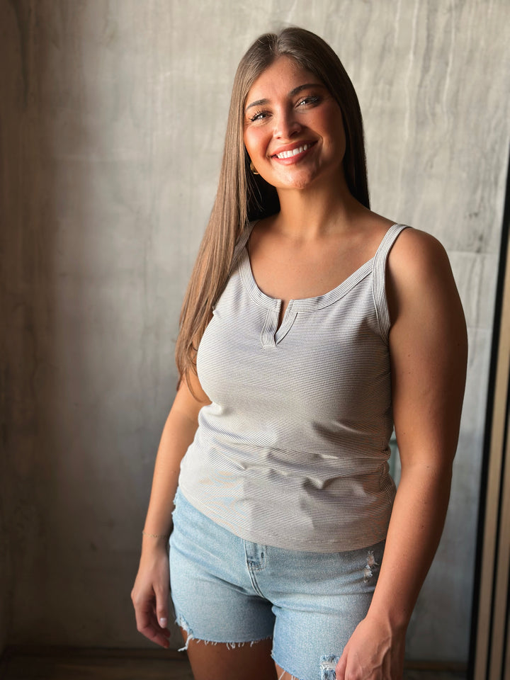 Woman wearing a light gray tank top and denim shorts standing against a neutral wall.