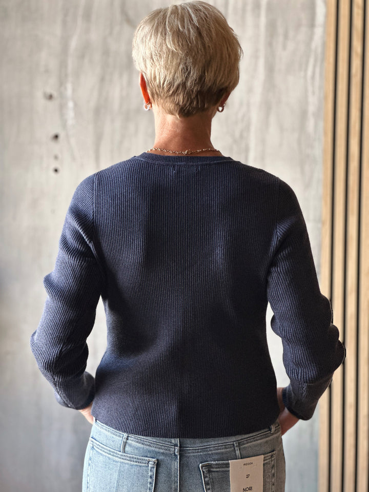 Person wearing a dark blue sweater and light blue jeans against a neutral wall.