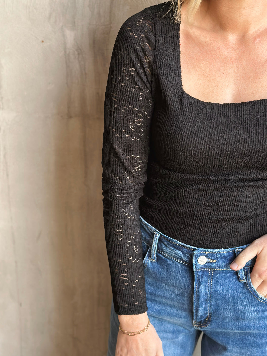 Person wearing a black long-sleeve top and blue jeans against a neutral wall.