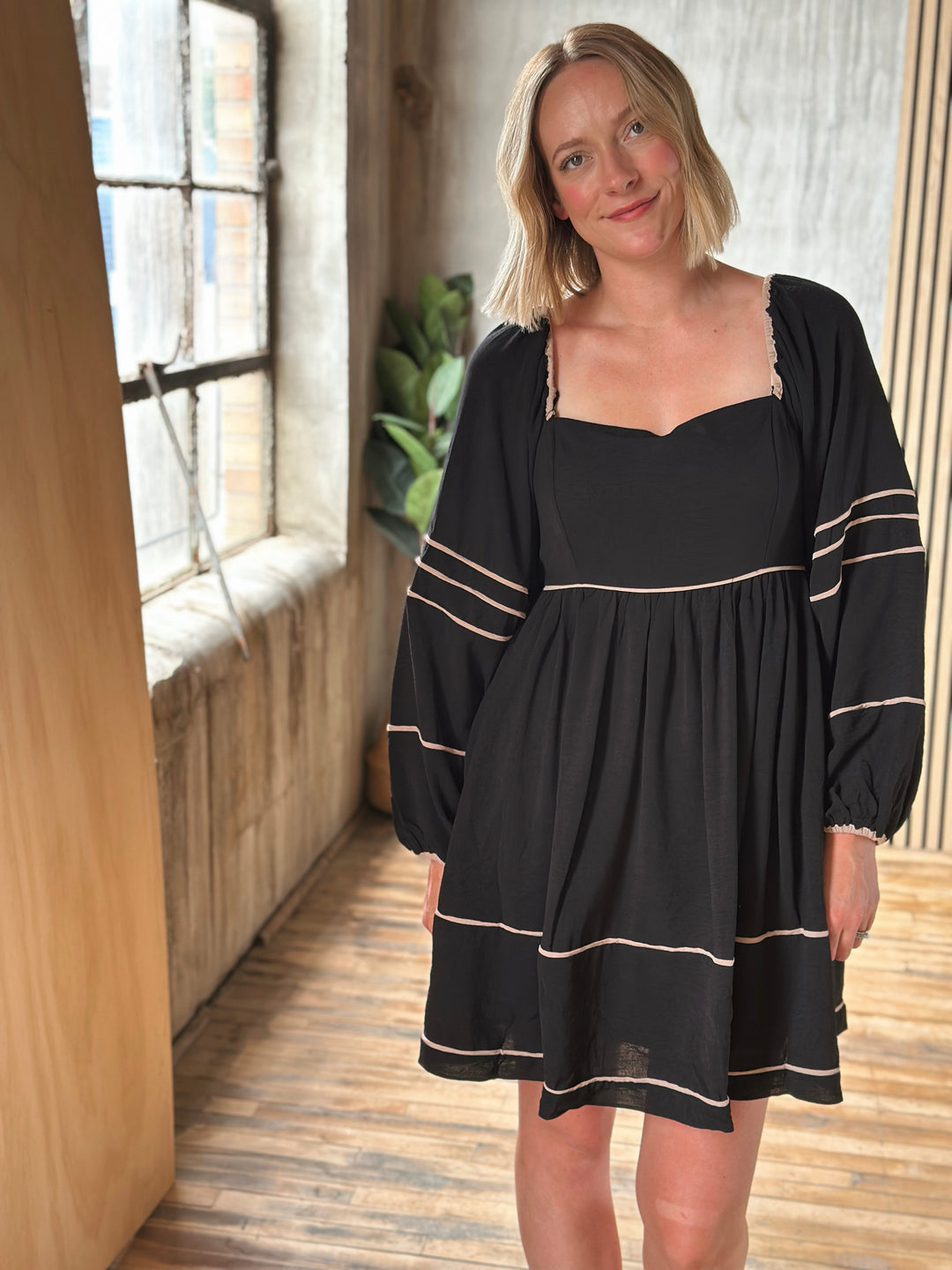 Woman wearing a black dress with long sleeves in a room with wooden floor and window.
