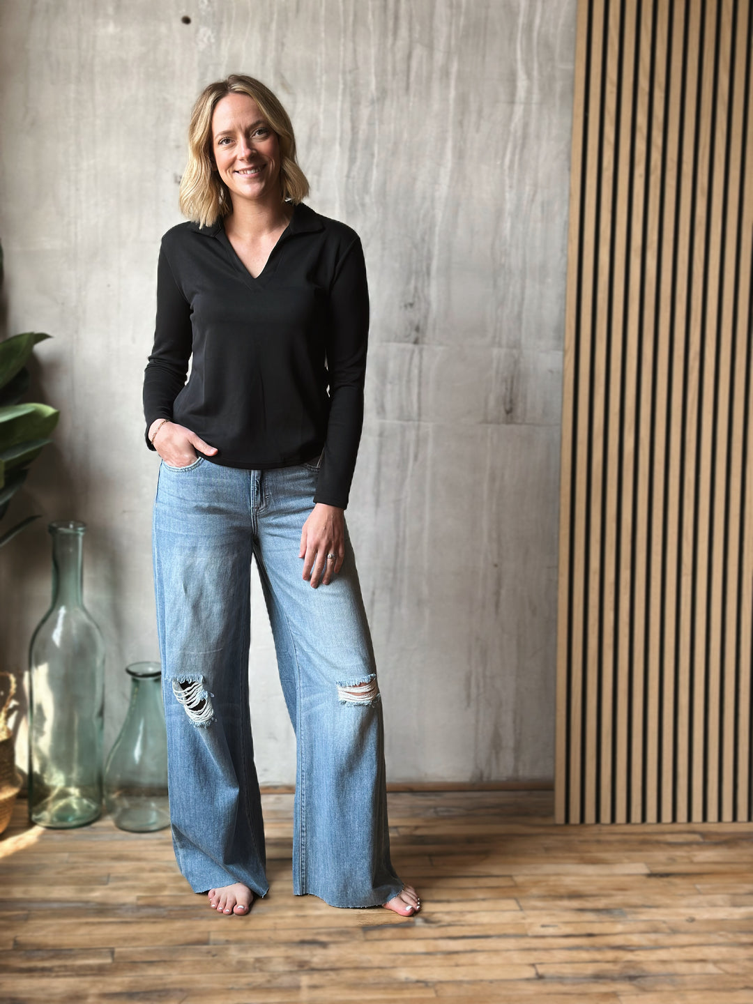 Woman wearing a black top and blue jeans standing in a room with a wooden floor and striped wall.