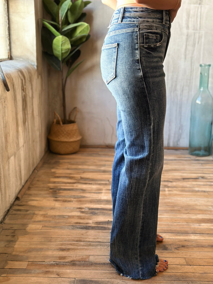 Person wearing blue jeans standing on a wooden floor with a plant and bottle in the background