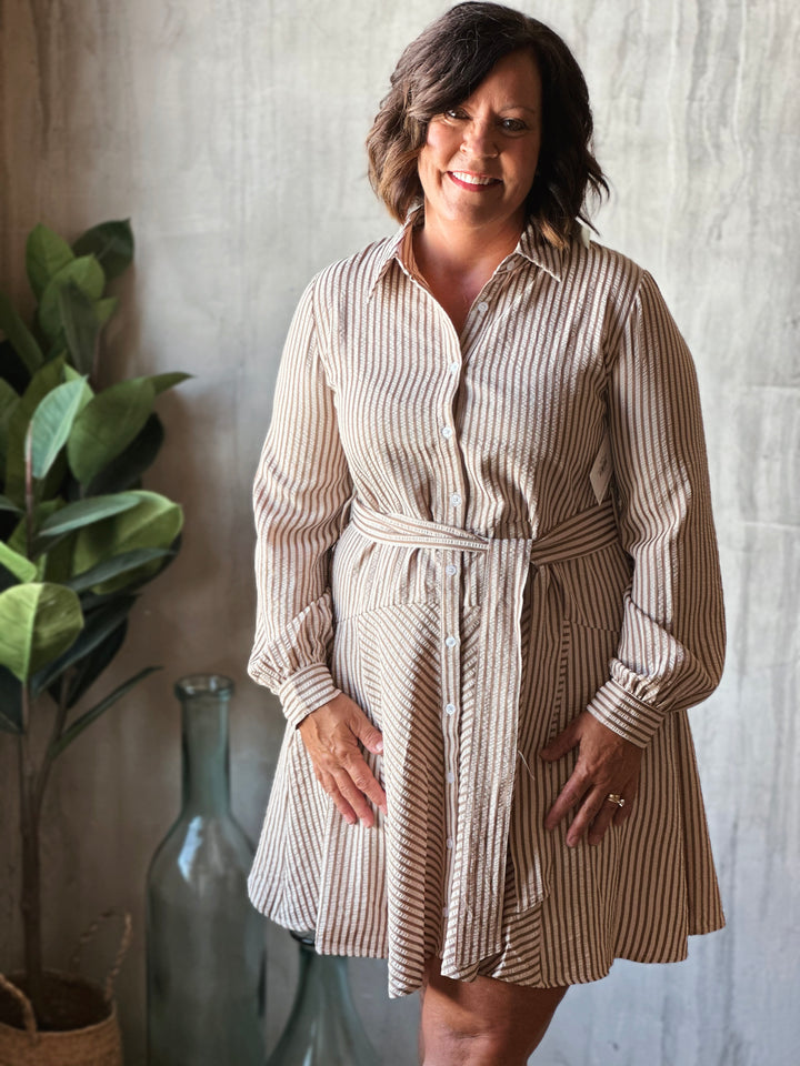 Woman wearing a striped dress standing against a neutral wall with a plant and vase in the background.