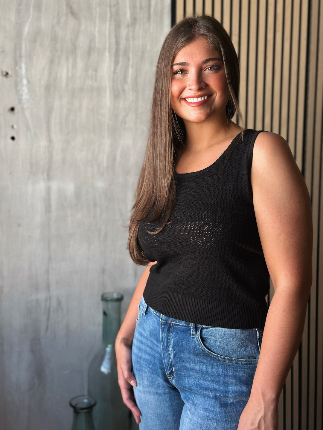 Woman wearing a black sleeveless top and blue jeans standing against a textured wall.