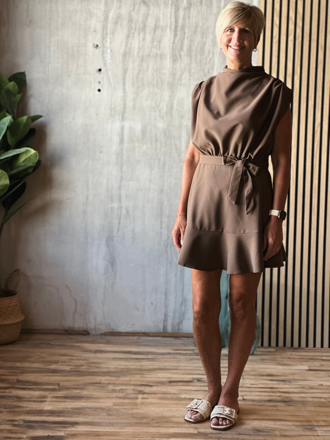 Woman wearing a brown dress standing indoors with a plant and striped wall in the background