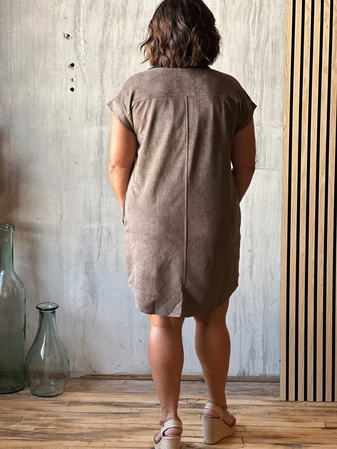 Person wearing a brown dress standing in a room with a concrete wall and wooden floor.