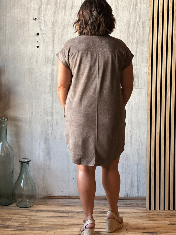 Person wearing a brown dress standing in a room with a concrete wall and wooden floor.