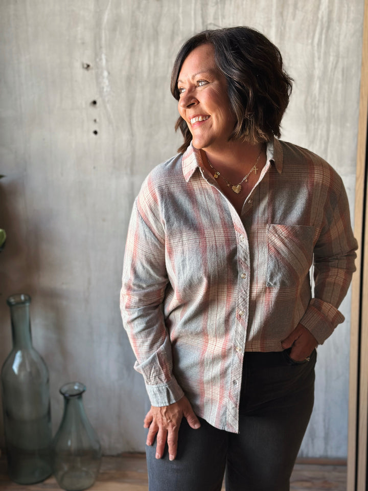 Woman wearing a plaid shirt standing against a textured wall with decorative bottles on a shelf.