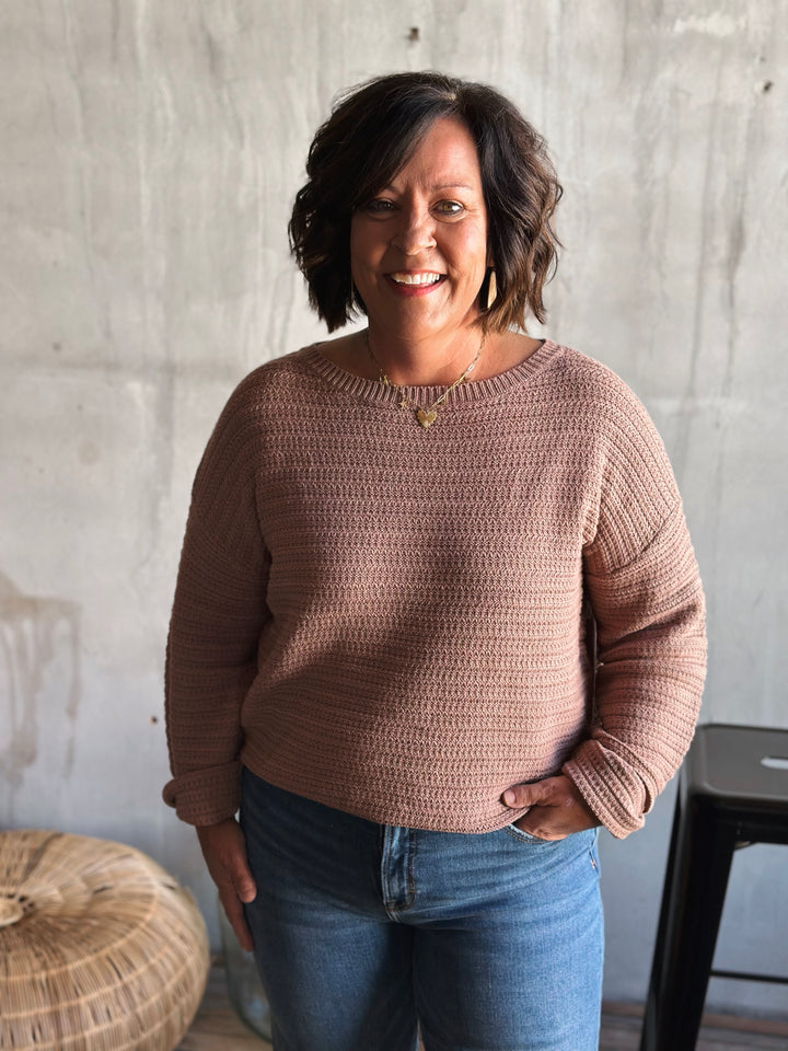 Woman wearing a brown crochet sweater and blue jeans standing against a concrete wall.