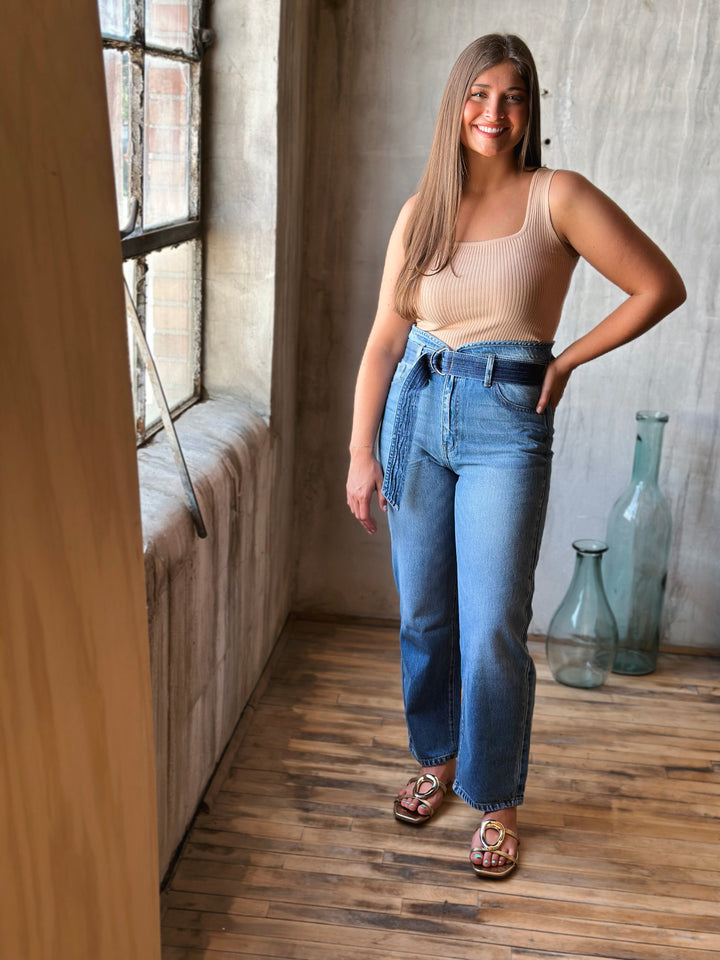 Woman wearing a beige tank top and blue jeans standing in a room with wooden floor and large window.