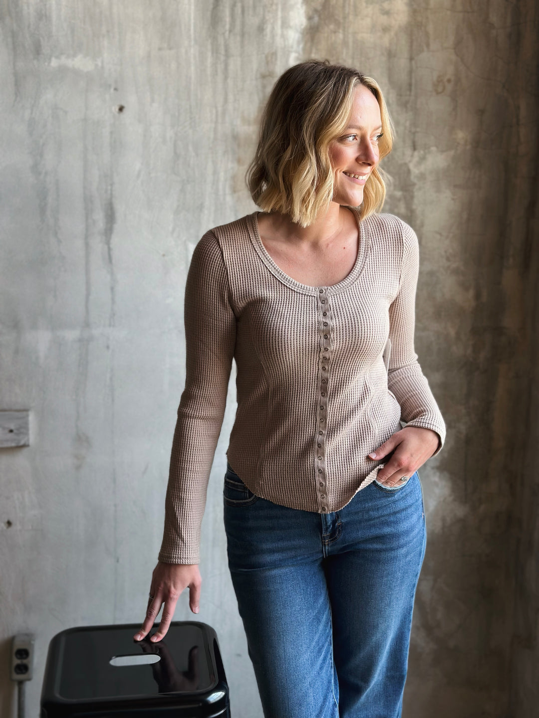 Woman wearing a beige long-sleeve shirt and blue jeans standing against a concrete wall.