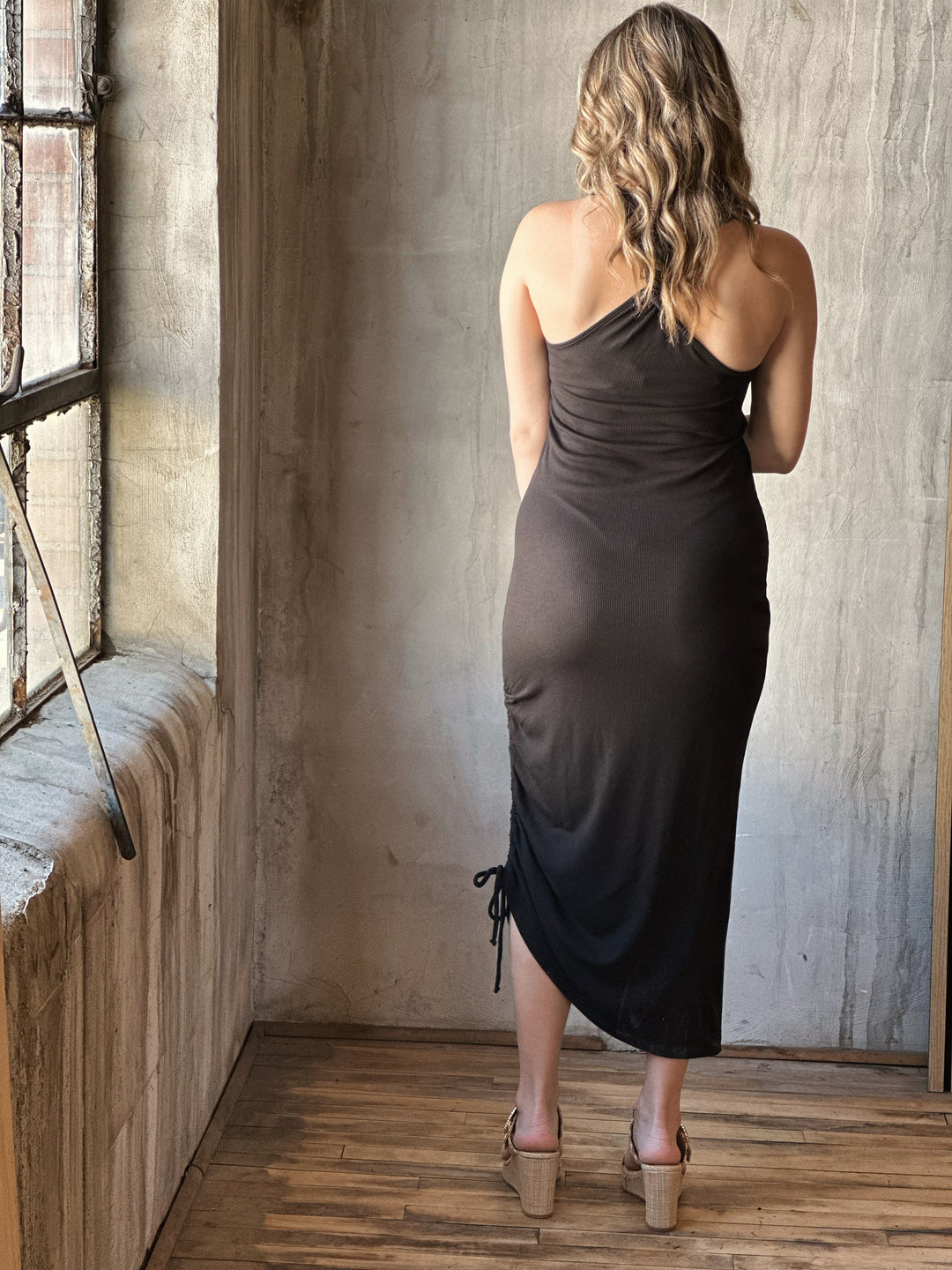 Back view of model wearing a black midi dress with a scoop neckline, sleeveless design and adjustable drawstrings along with a knotted open back detail