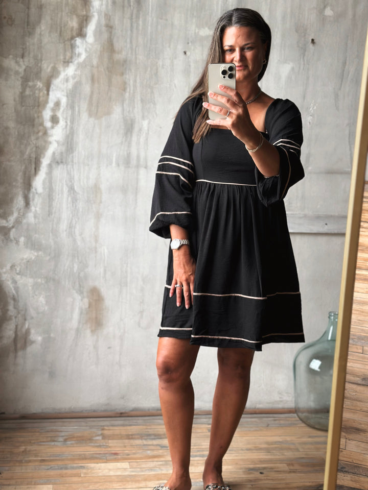 Woman taking a mirror selfie wearing a black dress with white trim.
