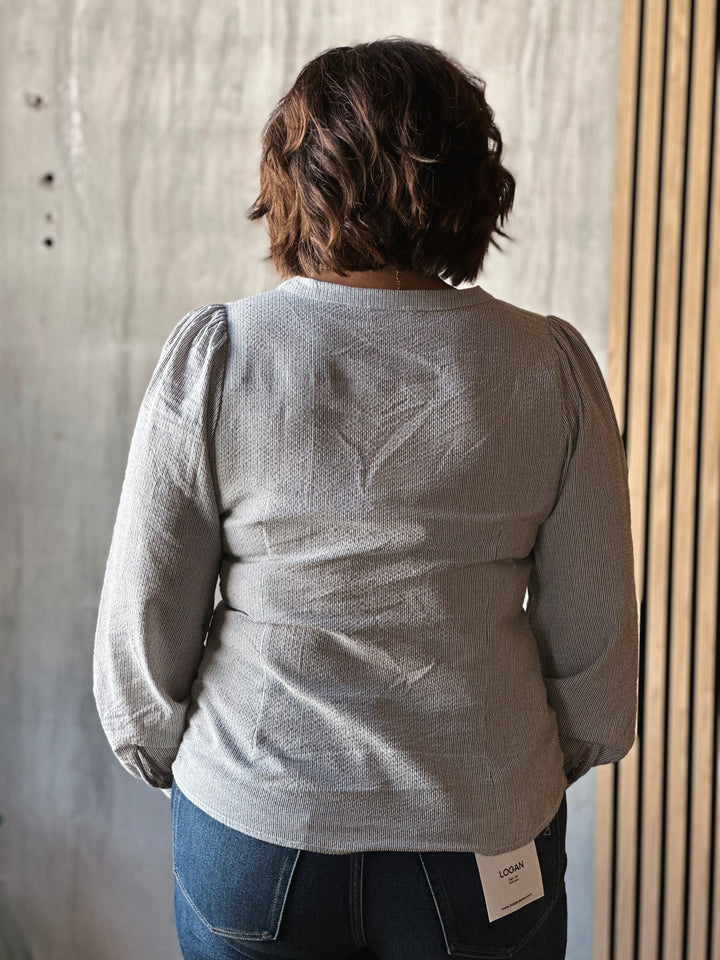 Person wearing a light gray textured sweater with a brand tag, standing against a neutral background.
