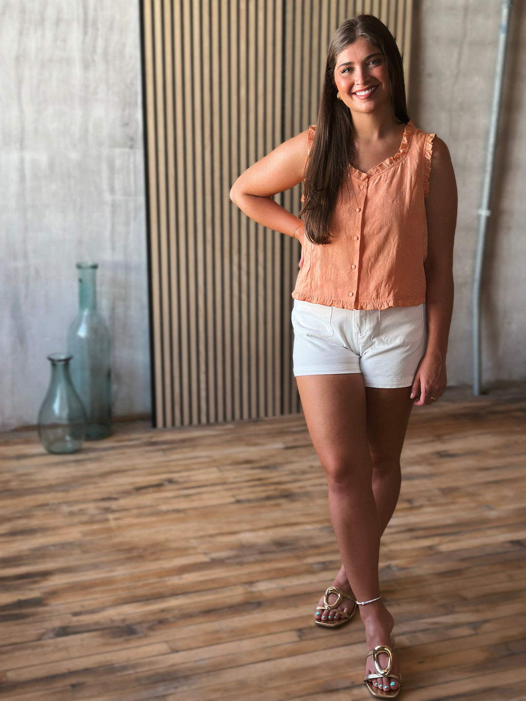 Woman in a peach blouse and white shorts standing in a room with wooden flooring and a textured wall.