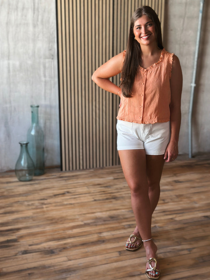 Woman in a peach blouse and white shorts standing in a room with wooden flooring and a textured wall.