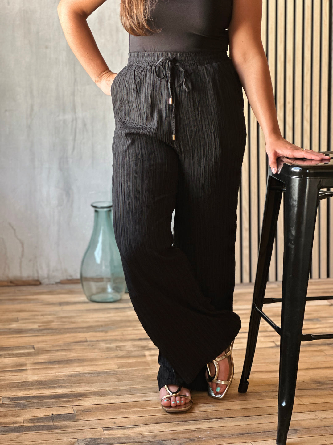 Person wearing black wide-leg pants standing next to a black stool indoors.