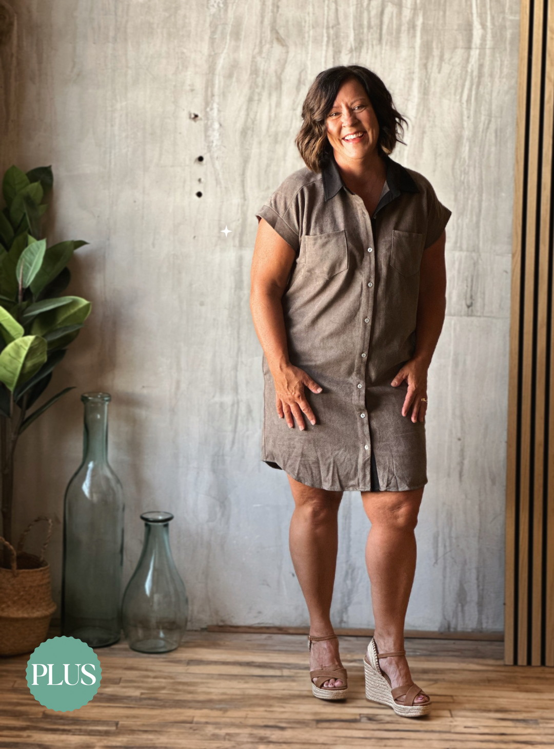 Woman wearing a brown dress standing against a wooden wall with decorative bottles and plants.