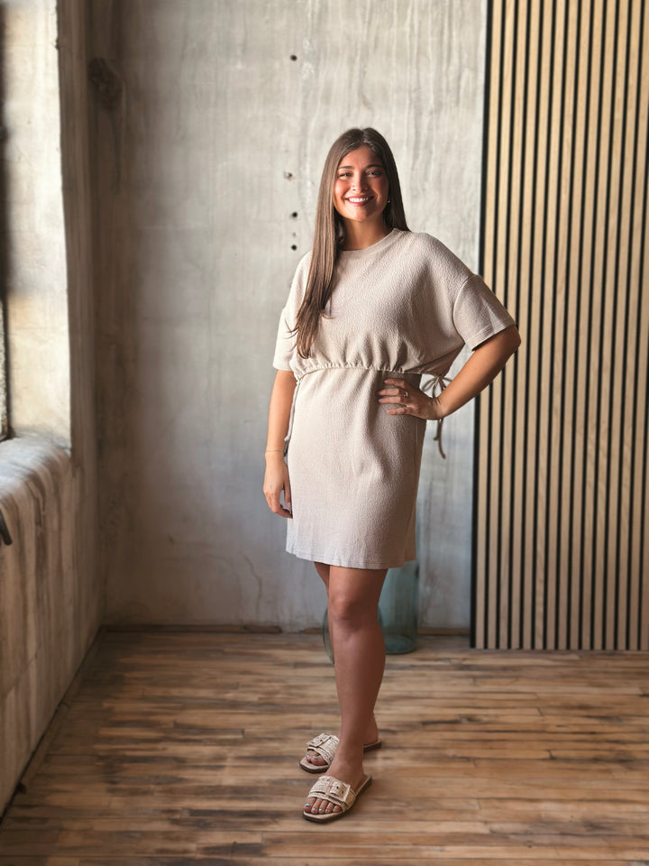 Woman in a beige dress standing in a room with wooden floor and concrete wall.