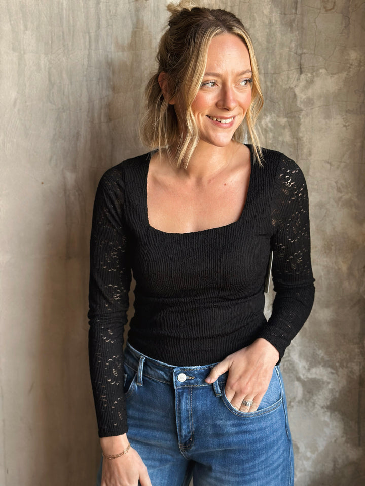 Woman wearing a black long-sleeve top and blue jeans against a textured wall.
