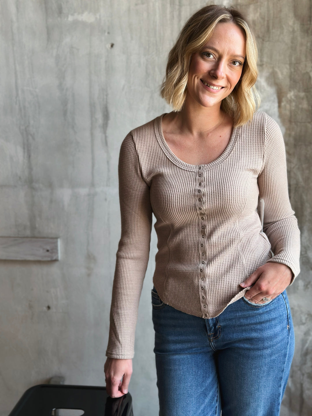 Woman wearing a beige long-sleeve shirt and blue jeans against a concrete wall.
