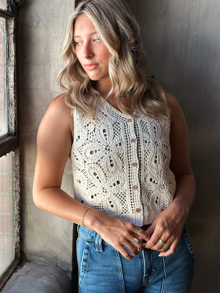 Woman wearing a white lace sleeveless top and blue jeans against a textured wall.
