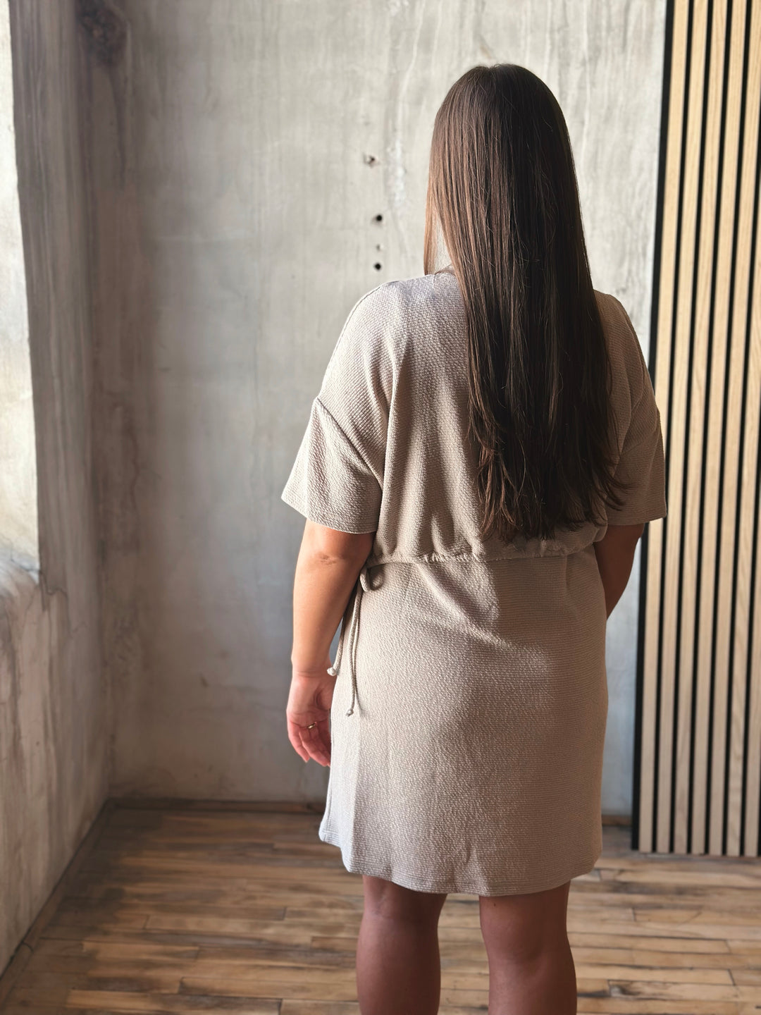 Person wearing a beige dress standing in a room with wooden floor and concrete wall.
