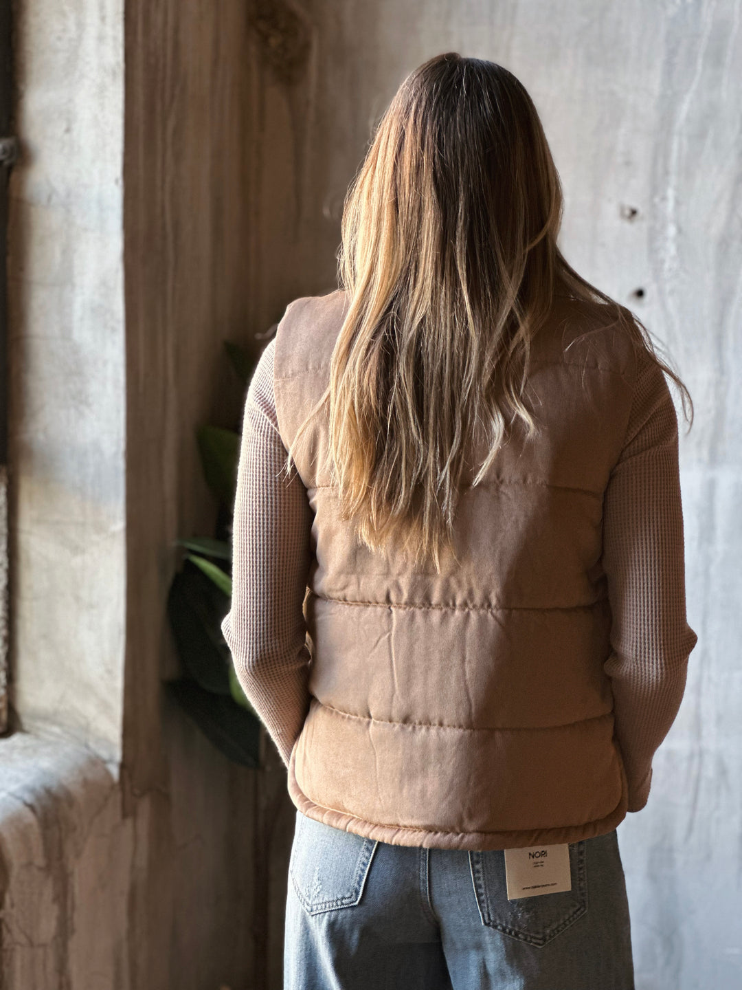 Person wearing a beige puffer vest and blue jeans standing in a rustic setting.
