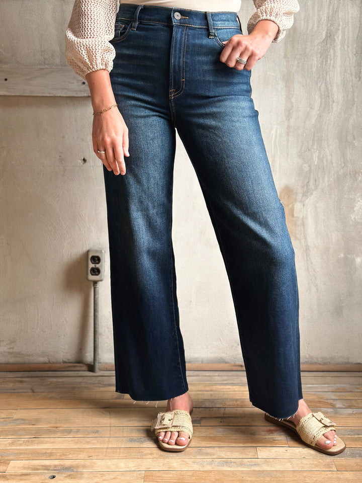 Person wearing blue jeans standing in a room with wooden flooring and a gray wall.
