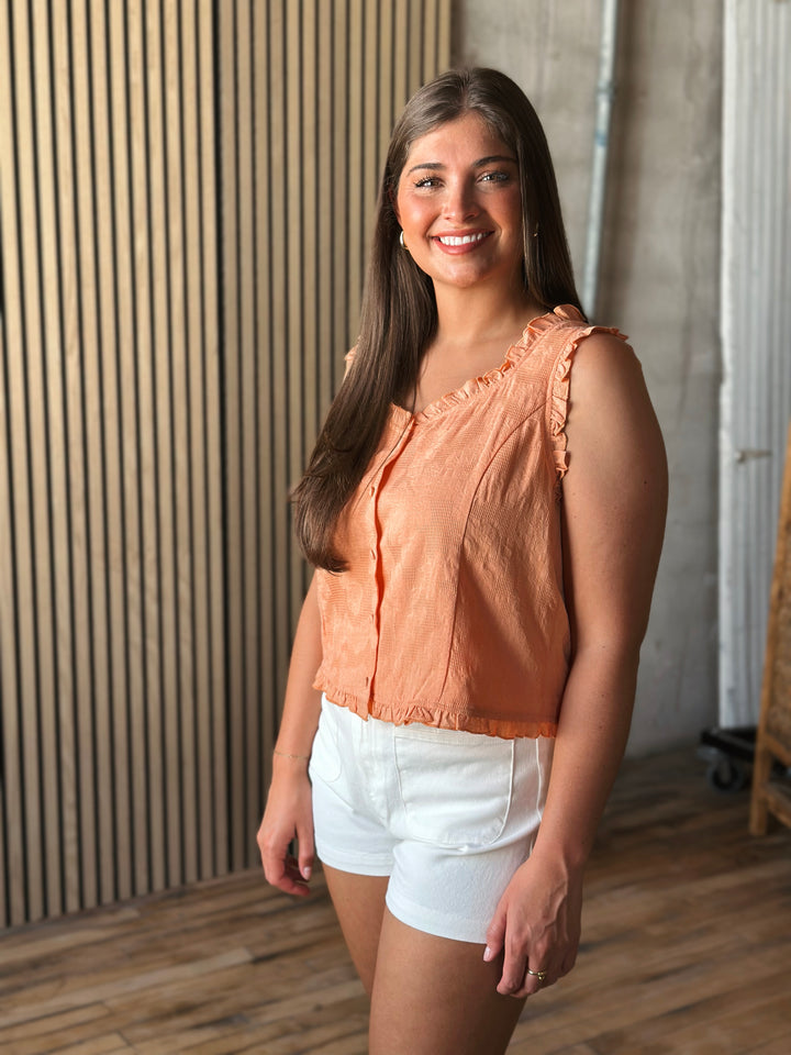 Woman wearing a peach sleeveless top and white shorts standing against a wooden paneled wall.