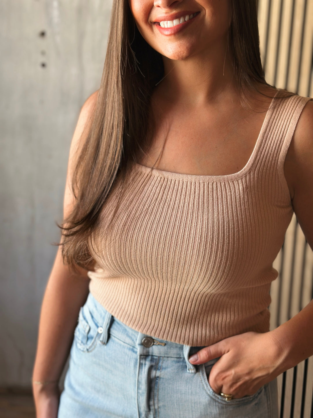 Woman wearing a beige ribbed tank top and light blue jeans indoors.