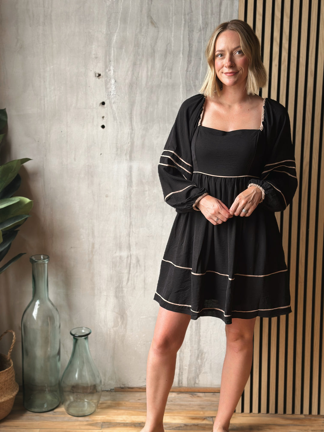 Woman wearing a black dress with white trim standing in a room with decorative vases and plants.